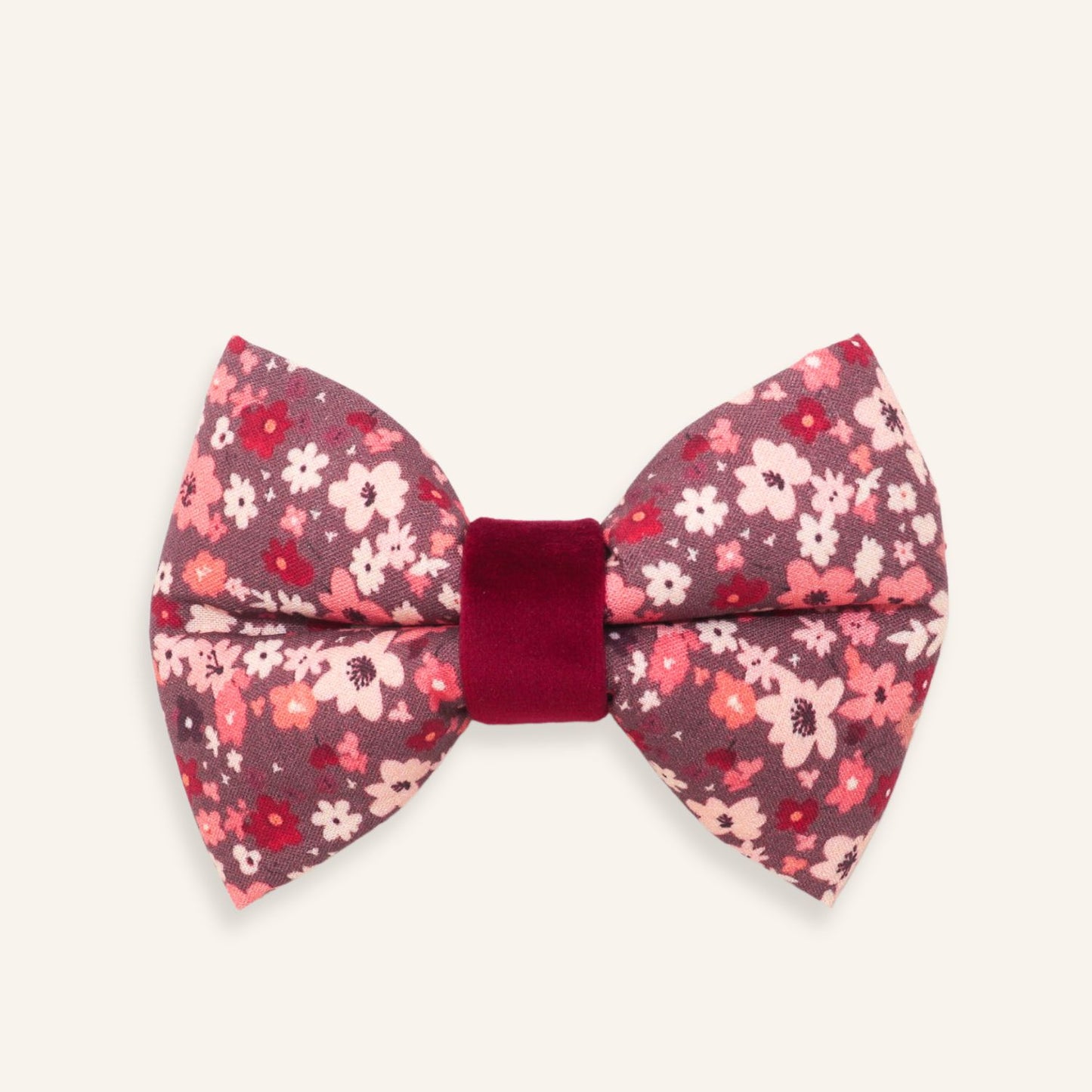 A small bow tie with a pink and red floral pattern on a dark mauve background, featuring a solid deep red velvet center band, shown on a light cream background.
