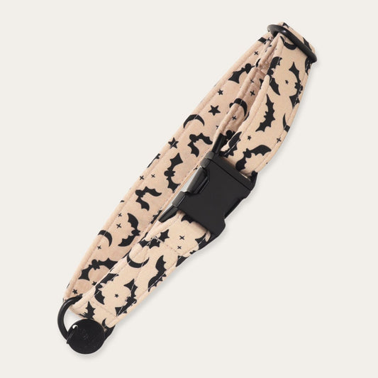 A fabric pet collar with a beige background printed with black bats, moons, and stars, featuring a black plastic quick-release buckle.