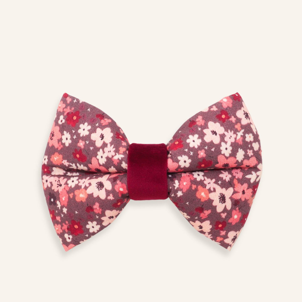 A small bow tie with a pink and red floral pattern on a dark mauve background, featuring a solid deep red velvet center band, shown on a light cream background.
