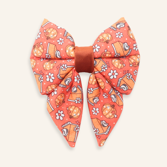 A large fabric hair bow featuring a warm orange and coral background with a pattern of pumpkins, candy corn, and white flowers, tied with a solid rust-brown center band, shown on a light cream background.