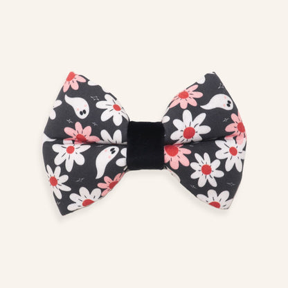 A black fabric bow tie featuring a cute Halloween pattern of white ghosts, white daisies with red centers, and pink daisies, set against a solid off-white background.