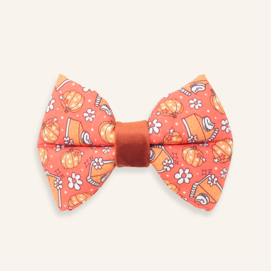 A fabric dog bow tie featuring a vibrant orange and coral background with a playful pattern of pumpkins, flowers, and autumn leaves, finished with a solid rust-brown center band.