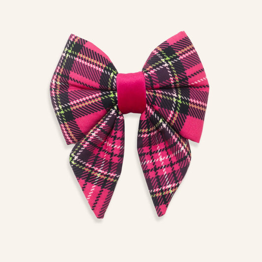 A large fabric bow featuring a vibrant pink and black tartan plaid pattern with thin white and green accent lines, set against a solid off-white background.